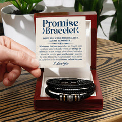 Hand holding a card with a heartfelt message above a black braided leather promise bracelet with metallic accents, displayed in a wooden gift box on a wooden table.