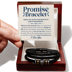 Hand holding a heartfelt message card above an open wooden box containing a black braided leather bracelet engraved with the words "love you" and "forever" designed as a sentimental gift.
