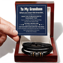 Hand holding a card with an inspirational message for a grandson, next to a braided black leather bracelet with metal accents, displayed inside a wooden gift box.