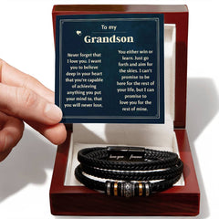 Hand holding a sentimental card with an engraved message for a grandson, displayed above a black braided leather bracelet inside a polished wooden gift box.