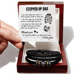 Hand holding a card with a heartfelt message for a stepdad above a dark brown wooden gift box containing a black braided leather bracelet with engraved metal accents.