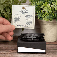 Black braided leather bracelet with a stainless steel cross resting on a gift box, accompanied by a graduation day message card for a special boy.