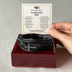 Hand holding a card with a heartfelt graduation message above a black braided leather bracelet featuring a stainless steel cross, displayed on a polished wooden gift box.