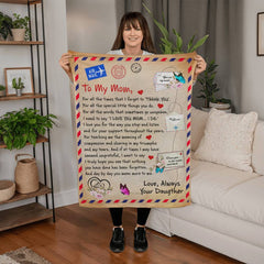 Woman holding a coral fleece blanket with a heartfelt letter design addressed to "Mom," decorated with hearts, butterflies, and postal stamp motifs in a cozy living room setting.