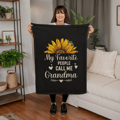 Woman holding a black coral fleece blanket with a large sunflower design and the phrase "My favorite people call me Grandma" in a cozy living room setting.