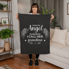 Woman holding a black coral fleece blanket with white angel wings and the heartfelt message "I have an angel in heaven I call her grandma," displayed in a cozy living room setting.
