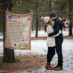 Couple dressed warmly in winter clothes embracing outdoors near a tree, with a heartfelt personalized coral fleece blanket hanging beside them in a snowy forest setting.