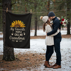 Black coral fleece blanket with sunflower design and the phrase "My Favorite People Call Me Grandma" hanging on a clothesline outdoors near a couple embracing in winter clothing.