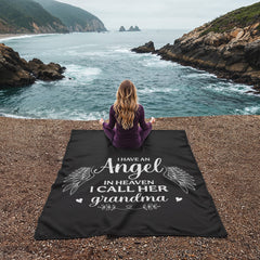 Woman sitting on a black coral fleece blanket with white script that reads "I have an Angel in Heaven I call her grandma," overlooking a rocky coastline and calm ocean.