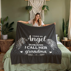 Woman holding a soft dark coral fleece blanket with white angel wings and the heartfelt phrase "I have an angel in heaven I call her grandma" in a cozy bedroom setting