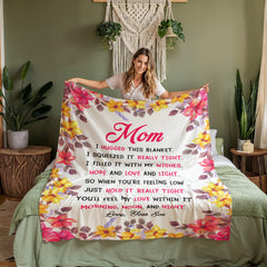 Woman holding a coral fleece blanket with a floral border and a heartfelt message from a son to his mom, displayed in a cozy bedroom setting with green walls and plants.
