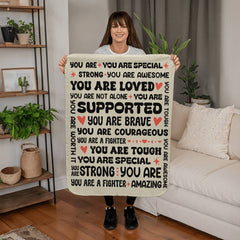 Woman holding a cream Sherpa fleece blanket with encouraging and uplifting phrases in black and red lettering in a cozy living room setting with plants and neutral decor.