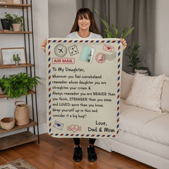 Woman holding a sherpa fleece blanket designed like an airmail letter with a heartfelt message from parents to their daughter, featuring decorative postage stamps and a cozy home setting.