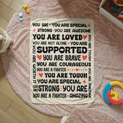 Cream-colored fleece blanket with encouraging and loving phrases like "You are loved," "You are brave," and "You are supported" in bold black and red text, displayed on a soft pink rug surrounded by children's toys.