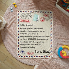 Sherpa fleece blanket designed like an airmail envelope with a heartfelt message from mom to daughter, placed on a soft pink rug surrounded by toys.