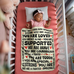 Baby girl sleeping peacefully in a crib, covered with a warm sherpa fleece blanket featuring encouraging and loving words in bold black and red text.