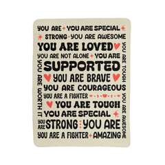 Cream-colored fleece blanket featuring positive, empowering affirmations in black and red text such as "You are loved," "You are brave," and "You are supported," designed to provide warmth and comfort.