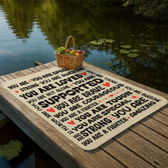 Cozy sherpa fleece blanket with inspirational phrases like "You are loved," "You are brave," and "You are amazing," laid out on a wooden dock beside a calm lake with a basket of fruit.