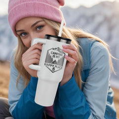 Young woman wearing a pink beanie and blue jacket holding a white insulated tumbler with a camping-themed design that reads "Camping is my happy place" while outdoors in a mountainous setting.