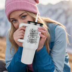 Young woman wearing a pink beanie and blue jacket holding a white laser engraved stainless steel tumbler with a humorous morning-themed message outdoors in a cozy setting.