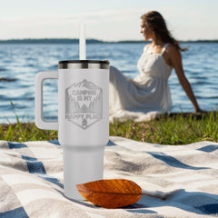 White insulated tumbler with a handle and black lid featuring a "Camping is my Happy Place" design, placed on a picnic blanket near a lake with a woman sitting in the background.