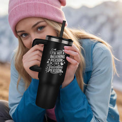 Young woman wearing a pink beanie and blue jacket holding a black stainless steel tumbler with a handle and straw, engraved with a humorous morning-themed message.