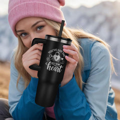 Young woman wearing a pink beanie and blue jacket holding a black insulated tumbler with a heart and key design and the phrase "You've got the key to my heart" outdoors in a mountainous setting.