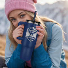 Young woman in a pink beanie and blue jacket holding a large navy blue insulated tumbler with a camping-themed design and a black straw outdoors in a mountain setting.