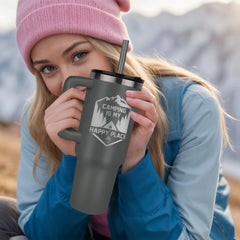 Young woman wearing a pink beanie and blue jacket holding a matte black tumbler engraved with "Camping is my happy place" outdoors in a mountainous setting.