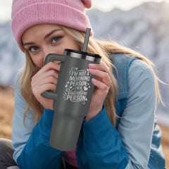 Young woman wearing a pink beanie and blue jacket holding a dark gray laser engraved tumbler with a humorous Valentine’s Day message outdoors in a mountainous setting.