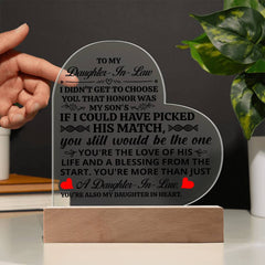 Hand holding a heart-shaped acrylic plaque with a heartfelt message to a daughter-in-law, displayed on a wooden base with a plant in the background.