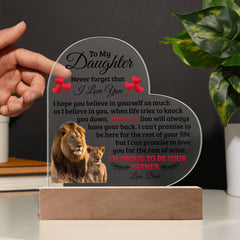 Hand holding a heart-shaped acrylic plaque with a heartfelt message from a father to his daughter, featuring an image of a lion and lion cub, displayed on a wooden base with a green plant in the background.