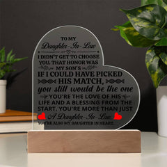 Heart-shaped acrylic plaque with a heartfelt message for a daughter-in-law, displayed on a wooden base, placed on a table next to green houseplants.