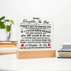 Clear acrylic plaque with heartfelt message for daughter-in-law, displayed on a wooden light-up base, placed on a white tabletop next to books and a small plant.