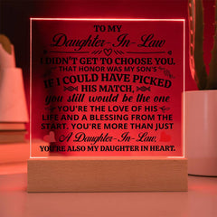 Illuminated acrylic plaque with heartfelt message for daughter-in-law, glowing with red LED light on a wooden base, displayed on a tabletop near plants.