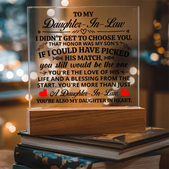 Clear acrylic plaque with an elegant message for a daughter-in-law, illuminated by a warm LED light on a wooden base, set on a stack of books with a cozy background.