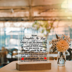 Clear acrylic plaque with a heartfelt message for a daughter-in-law, illuminated by a glowing LED base, displayed next to a small vase of flowers on a wooden table.