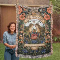 Decorative woven heirloom blanket featuring two frogs facing each other with floral patterns and the phrase "Until We Croak," displayed outdoors with a person in the background.