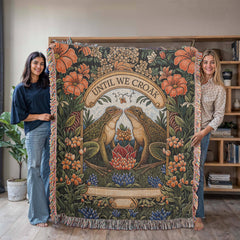 Two women holding a woven heirloom cotton blanket featuring detailed floral patterns and two frogs facing each other beneath a banner reading "Until We Croak," in a cozy indoor setting.
