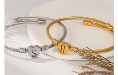 Two textured cable bracelets in silver and rose gold, each featuring a personalized heart-shaped initial charm, displayed on a white surface with dried flowers nearby.