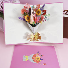 Open 3D pop-up card featuring a colorful floral bouquet with sunflowers and a mother holding a child, placed on a pink surface with a matching envelope showing a floral illustration.