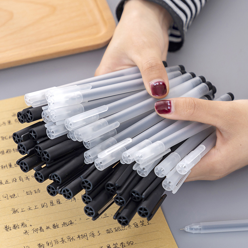Hand holding a bundle of pens with black barrels and translucent caps, resting over a sheet of handwritten text on a wooden surface.