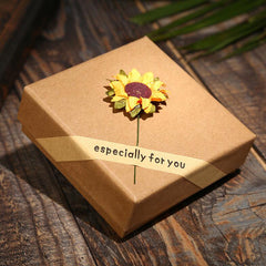 Brown gift box with a small sunflower decoration and a beige ribbon that reads "especially for you," placed on a wooden surface in soft natural light.