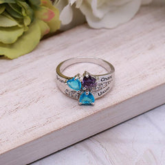 Silver personalized ring with three heart-shaped birthstones in blue and purple, custom engraved with family names, displayed on a wooden surface.