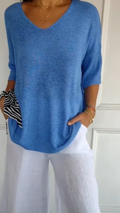 Woman wearing a stretchable blue V-neck knit sweater with half sleeves paired with white linen pants, accessorized with gold layered necklaces and bracelets, holding a striped fabric.