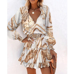 Woman wearing a stylish long sleeve patchwork dress with puff sleeves and a ruffled hem in neutral tones, accessorized with layered gold necklaces and holding a brown clutch.