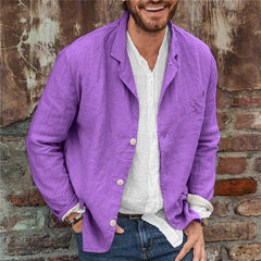 Man wearing a slim fit casual sport coat in purple cotton linen over a white shirt and jeans, standing against a rustic brick wall background.