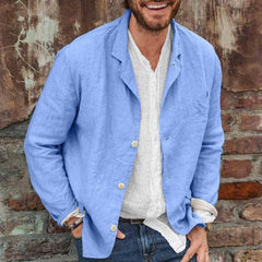 Man wearing a slim fit casual sport coat made from lightweight cotton linen in light blue over a white shirt, standing against a rustic brick and textured wall background.