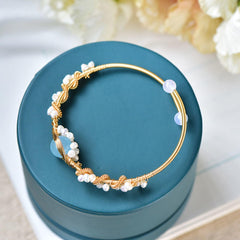Gold wire braided bracelet adorned with delicate white pearls displayed on a teal round jewelry box, set against a soft floral background.
